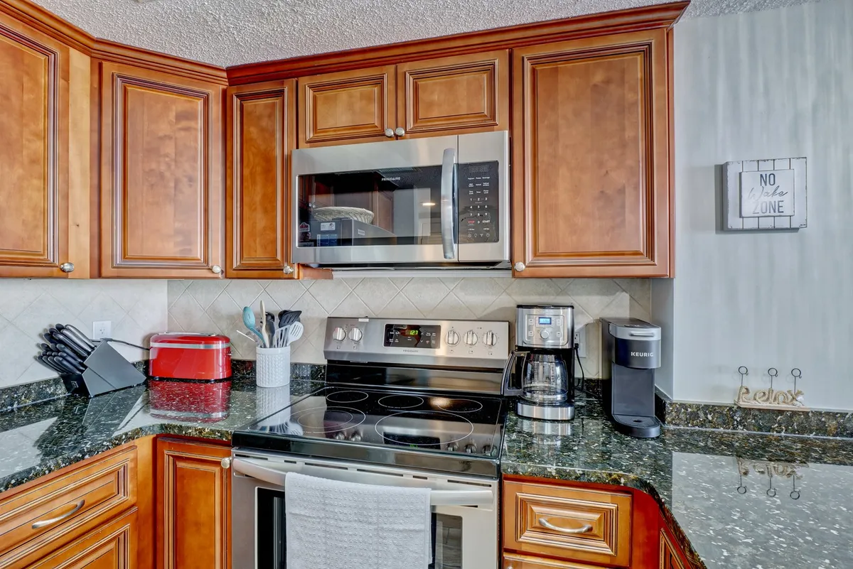A bright, modern kitchen is equipped with new stainless steel appliances, gleaming granite countertops, ample cabinetry, a coffee maker, and a cozy decorative sign. The layout promotes easy meal prep and informal entertaining, while the design connects seamlessly to adjacent gathering spaces.