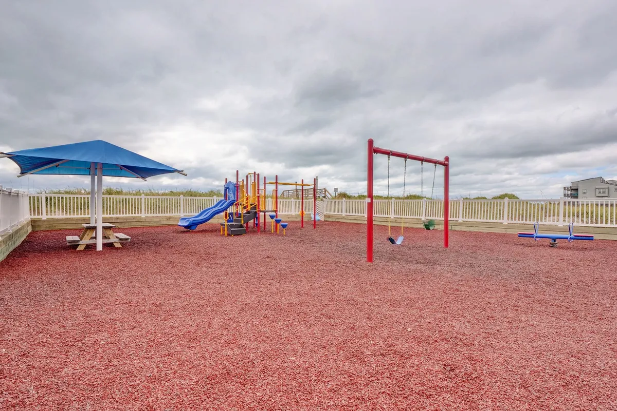 The image captures an inviting playground scene with swings, play structures, a picnic table, and an umbrella, set in a communal outdoor area. Children are seen enjoying playtime while parents relax nearby.