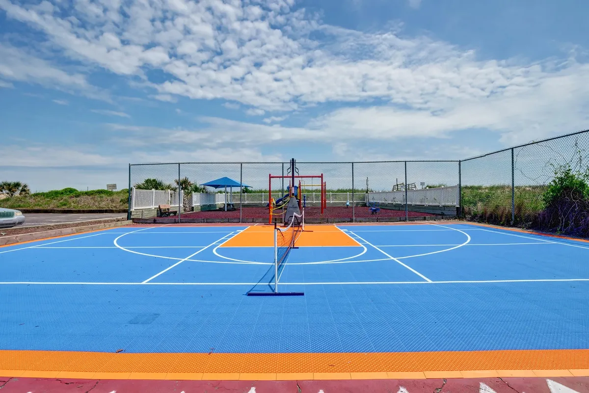 A vibrant, colorful court is depicted with a basketball hoop and net, surrounded by greenery. The area invites playful activities and family competitions, suggesting a place for basketball, tennis, or pickleball.