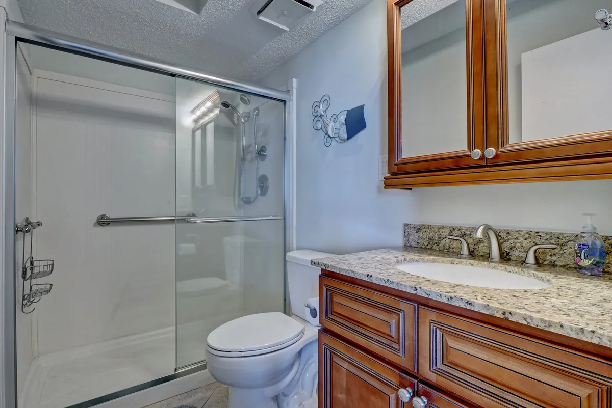 A modern ensuite bath is adorned with a sleek granite countertop, wooden cabinetry, a glass shower enclosure, and stylish fixtures. The space is bright and organized, inviting guests to unwind in the walk-in shower after a day at the beach.