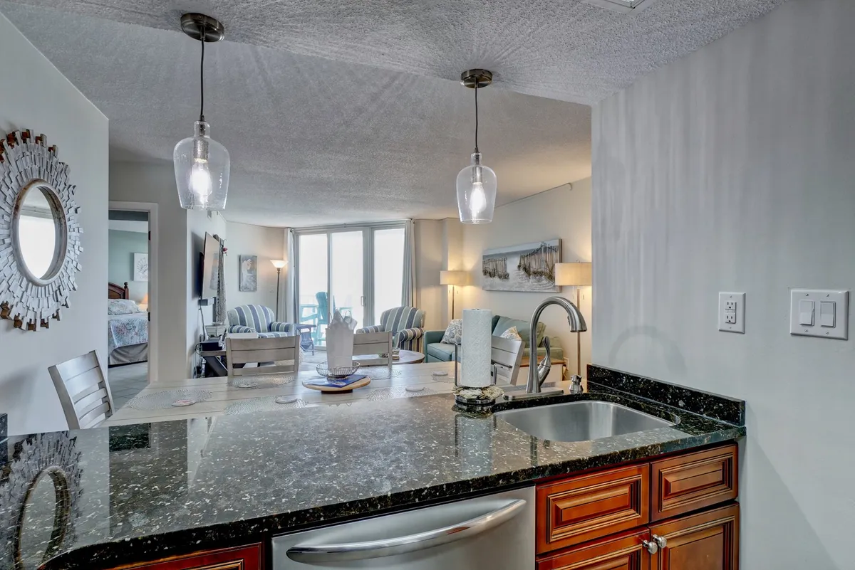 This inviting open-concept living and dining area features a spacious granite countertop, modern pendant lighting, a large dining table set for gatherings, and a comfortable living area with plush seating and decorative lamps. The overall ambiance is warm and welcoming, perfect for socializing and enjoying ocean views through expansive windows (not pictured directly, but strongly implied by the layout and lighting).