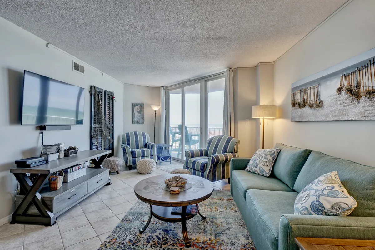 A comfortable living room furnished with a plush green sofa, wooden coffee table, striped accent chairs, a large TV, and decorative wall art. The setup invites relaxation, movie nights, and game sessions, with natural light hinting at ocean views beyond.