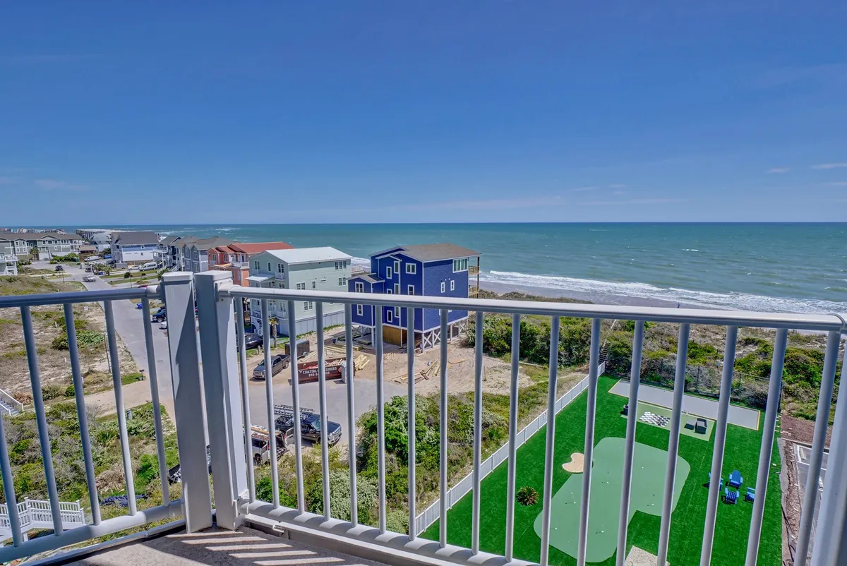 This image features an inviting balcony overlooking expansive green lawns and the vibrant Atlantic coastline, framed by colorful houses. The setting is serene, with people relaxing on the balcony and enjoying the fresh ocean breezes, evoking the sense of leisurely mornings or evenings spent together.