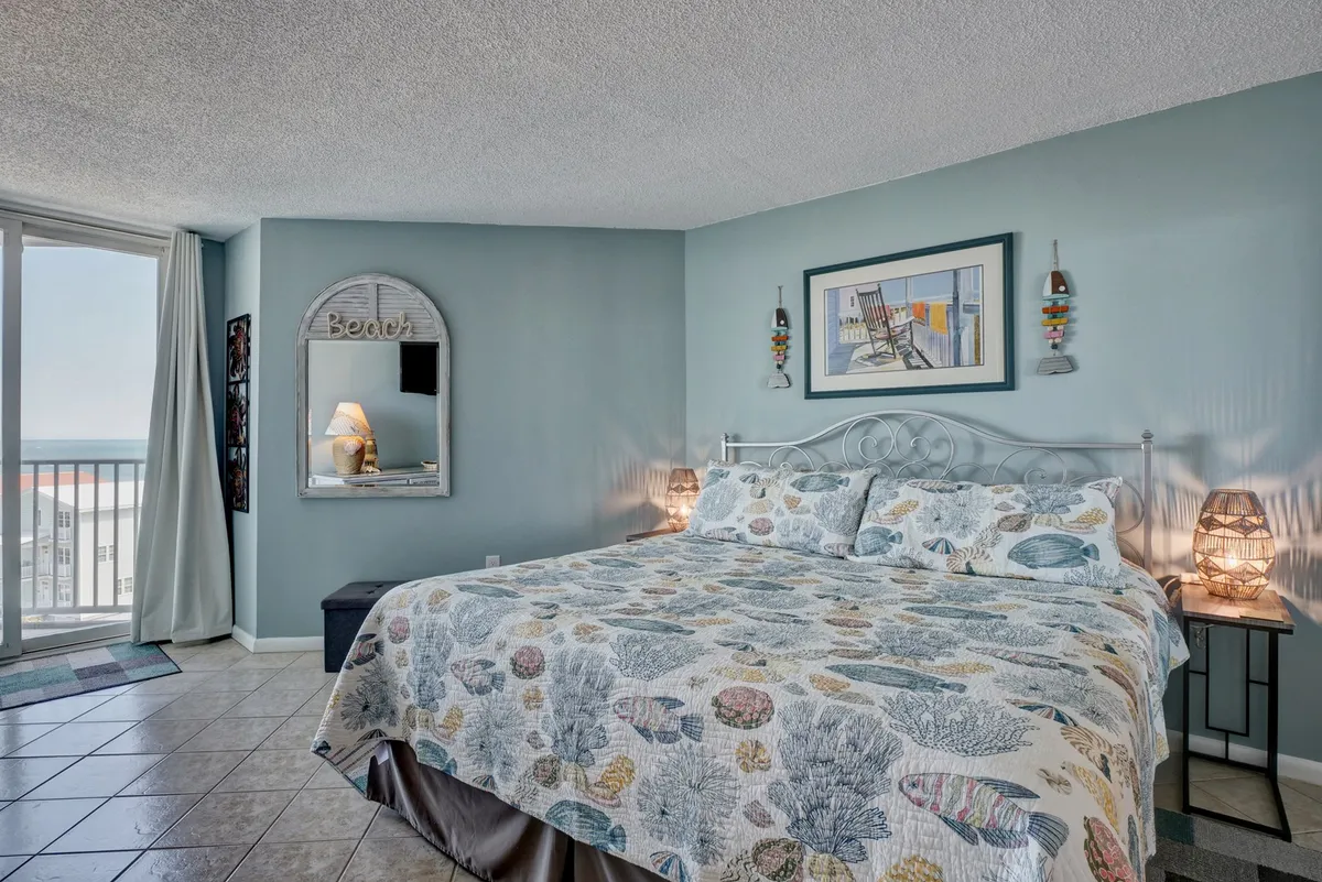 This image features a cozy bed with a patterned quilt situated next to a large window that offers sweeping ocean views. Decorative art and a mirror add a touch of elegance, while natural light fills the room, creating a calm, tranquil atmosphere ideal for relaxation or waking up to the coastal scenery.