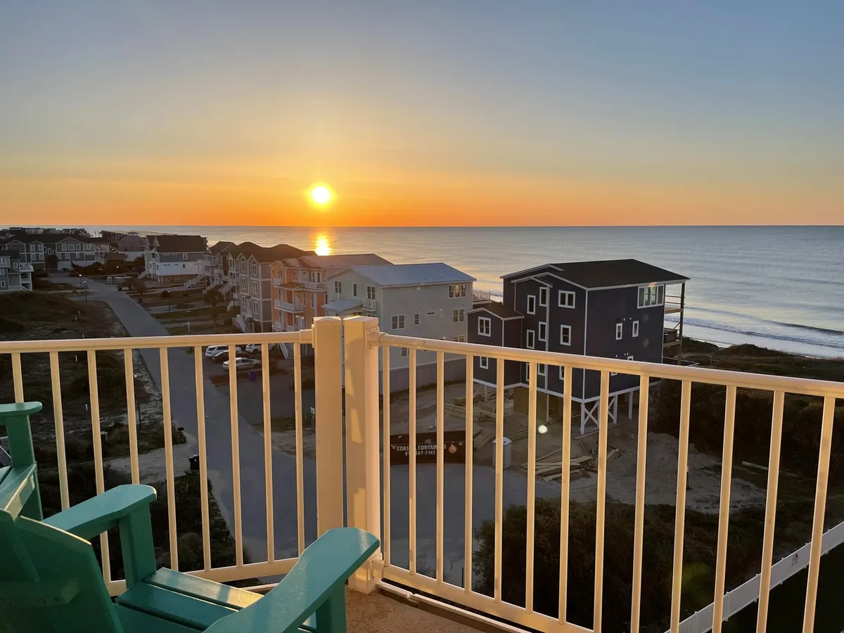 The image captures a peaceful scene from the balcony of a North Topsail Beach condo. Two chairs sit invitingly behind a railing, overlooking a breathtaking sunset as golden hues reflect over the tranquil ocean. In the distance, beach houses dot the shore. The mood is serene, emphasizing quiet relaxation and the beauty of the natural surroundings.