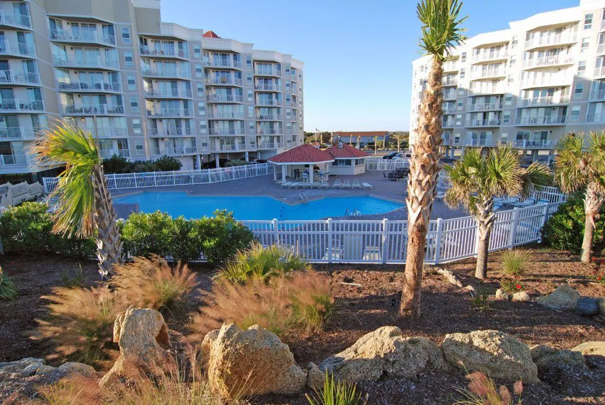 This image showcases a sparkling swimming pool surrounded by lush greenery and palm trees, with residential resort buildings in the background. People are seen swimming, sunbathing, and relaxing by the pool, creating a lively yet serene resort atmosphere.