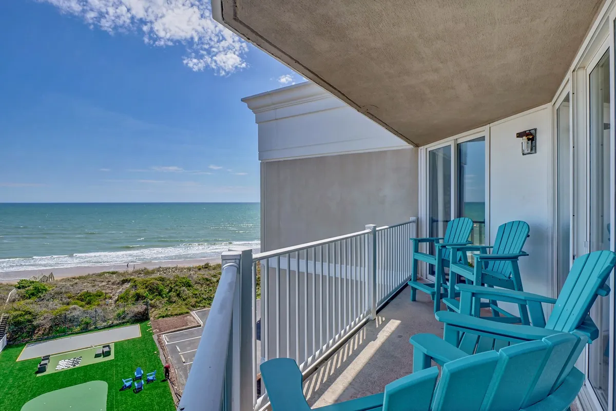 A tranquil balcony scene with inviting blue chairs offers an uninterrupted view of the ocean and a lush green lawn below. The space feels tailored for quiet moments, stargazing, or shared drinks, with the gentle sound of waves in the background.