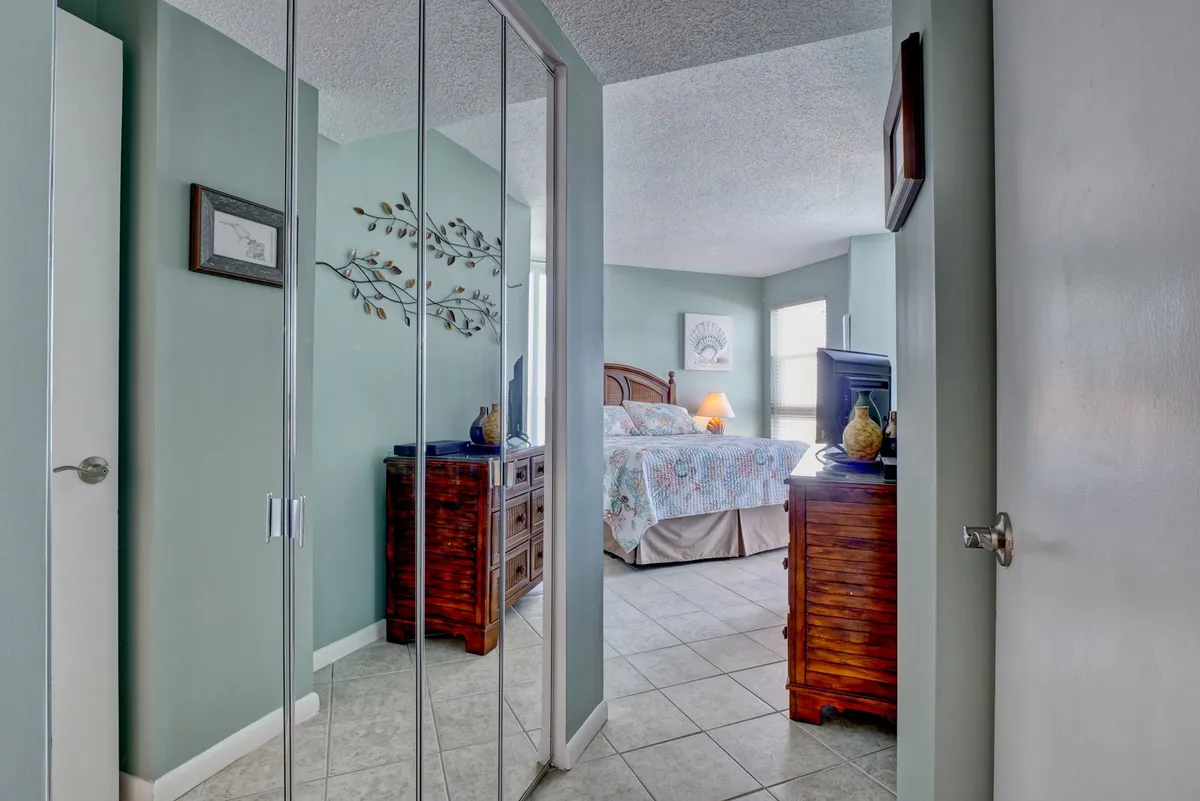 The bedroom is styled with mirrored closet doors, wooden nightstands, plush bedding with floral patterns, and framed wall art. Gentle morning light streams in, highlighting the restful and inviting atmosphere, perfect for winding down or starting the day refreshed.
