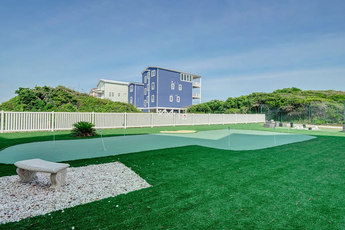 This exterior image showcases a manicured putting green in front of a blue house, complete with green turf, a decorative stone bench, and lush landscaping. The atmosphere is relaxed and inviting, ideal for morning golf sessions or unwinding outdoors.