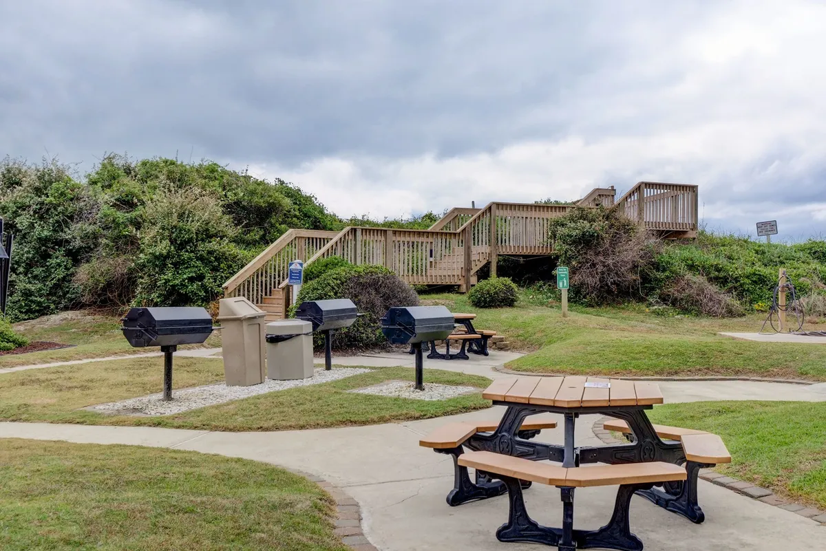 An outdoor communal grilling area with barbecue grills, a picnic table, wooden stairs, and surrounding greenery sets the scene for relaxed social gatherings, meals, and connection with nature.
